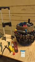 Photo showing the bucket organizer with many screwdrivers in exterior pockets, tool dolly frame with clip-on wheels beside it, and some tools laid out on a wooden surface including pliers, levels, and measuring tools.