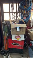 Red and black Lincoln arc welder with hoses and green cylinder nearby, cardboard box on top