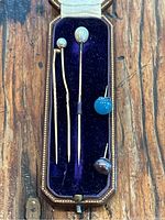 All items shown arranged inside original brown leather presentation box with blue velvet insert and cream silk lining, displaying three gold stick pins topped with pearls and two loose earrings, one blue stone detached, one dark pearl earring.