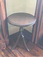 Front angle view of antique style wooden round pedestal table showing top, pedestal column and three curved legs with carved edge detail on table top