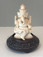 Front view of ivory-colored carved Ganesha figure on dark carved wooden circular base, showing broken right hand.
