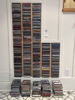 Wooden wall-mounted rack holding six columns of compact discs in jewel cases and additional stacks on floor
