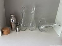 Front view of all barware items including 3 glass decanters with stoppers, chrome martini shaker, shot measure, and cork coasters on white shelf.
