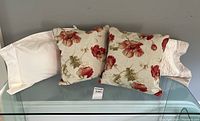 Four decorative throw pillows arranged on a glass table against a gray wall. Two have a red poppy floral print on linen, two are cream silk with a quilted pattern.