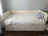 White IKEA single bed with beadboard paneling on the backrest and side resting walls. Three storage drawers are built into the base of the bed, each with a round black knob. Bed is made of wood with white painted finish. Bedding includes white and gray pillows and a white striped duvet. Minor repairs made to one side of the bed base.