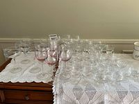 Wide view of the lot showing multiple clear coupe style glasses, smaller and larger clear stemmed wine glasses, and pink stemmed wine glasses arranged on lace tablecloth.