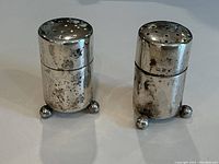 Front angle of two cylindrical sterling silver shakers with visible tarnish, placed upright on ball feet.