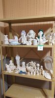 Wide view of three shelves filled with various unpainted and partially painted ceramic figurines including human figures, animals, hats, and decorative pottery pieces.