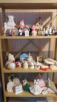Ceramic figures and pieces arranged on a 3-tier beige metal shelf, showing painted and unpainted figurines including animal, human, and decorative forms.