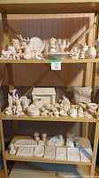 Photo showing three shelves of unglazed ceramic pieces including animals, human figurines, plaques, and small containers, arranged on a wooden shelving unit.