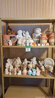 Shelf holding a variety of painted and unpainted ceramic items including figurines, jars, and decorative pieces arranged on two shelves.