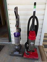 Side view of two upright vacuum cleaners: left Dyson DC42 with transparent bin and purple cyclone, right Dirt Devil Featherlite in red and grey with hose attached.