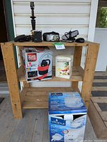 Wooden shelf with various electronics and household items including boxed wet & dry vacuum, boxed Vitapur filtration system, camera, speaker, alarm clock, mouse, and lamp base.
