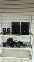 Wide shot showing all items on shelving: 4 speakers, 2 cameras, camera bag and accessories, manuals, cords.