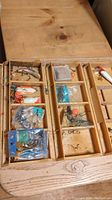 Top view of foldable wooden tackle box opened flat on table showing multiple small fishing lures, hooks, and terminal tackle items in compartments.