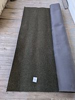 Rolled out section of dark forest green commercial carpet on wooden floor, showing clean top surface and gray backing partially rolled up.