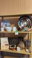 Photo of two shelves showing metal mixing bowl, Pyrex mixing bowls, vintage Betty Crocker recipe box, mugs, baking pans, blender pitcher, and pot holders