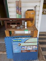 Photo showing vintage Canada Dry wooden bottle holder crate, rolled cork mats, yellow jug, curtain rods, clear globe decorations, small white spray bottle, cushion mat, and wooden stand on a wooden bench.