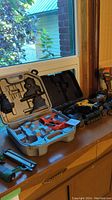 Photo of assorted power tools on a wooden surface near window, includes cordless drill, power nail guns, and an orange power tool in a case.