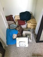 Stack of various folded bed linens laid on floor near closet, including sheets, blankets, cushions, and bed skirts in multiple colors.