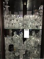 Photo showing multiple cut glass decanters, stemware glasses, and candy bowls on two glass shelves inside a dark wood cabinet.