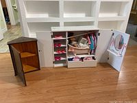 White doll closet case open, showing shelves with pink doll shoes, doll bathtub with doll inside, several doll clothes on hangers, and drawer open with doll clothes.