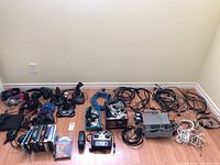 Wide shot of gaming controllers, game discs, headphones, Blu-ray players, and cables arranged on floor.