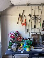 Photo showing assorted garden tools hanging on wall including hedge trimmer, pruners, a spray wand. Watering cans, garden accessories, and decorative flowers on table underneath.