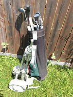 Full view of golf clubs in bag with pull cart leaning against fence in grass.