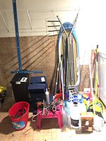 Photo showing ironing board, folding storage boxes, brooms, mops, red bucket, cleaning products, pink step stool, and black garbage pail.