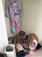 Wide view of tie-dye unicorn onesie hanging on wall above pile of toys, books, and dress-up items scattered on floor including hats and jewelry