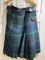 Traditional pleated Scottish tartan wool kilt with black leather belt and buckles hanging on a hanger.