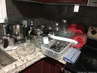 Overview of assorted kitchen items including Corning Ware glass dishes with lids, Lagostina pots with lids, Sunbeam kettle, and a plastic container with cutlery.