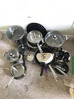 Photo showing various pots, pans, and strainers arranged on carpet including large stainless steel pots and frying pans
