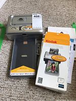 Silver Kodak EasyShare printer dock plus with original box and manuals, front paper tray extended, showing Kodak logo on the tray.