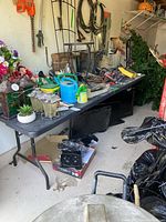 Black plastic folding table holding various gardening tools and miscellaneous items, located in a garage or storage area, showing dirt and signs of wear