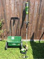 Photo showing both the Scotts seed spreader and Greenworks string trimmer leaning against wooden fence on grass.