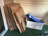 Flattened medium and large cardboard moving boxes stacked vertically against exterior wall next to a large white plastic storage tote with an off-white lid and blue packing straps inside.