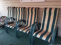 Four rigid green metal patio chairs with green and beige striped floral fabric seats and backs. Chairs are lined up against siding on porch.