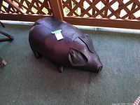 Photo of brown leather pig-shaped ottoman with visible stitching, ears, snout and legs, placed on green outdoor carpet near lattice wood rail.