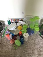 Photo shows assorted plastic storage containers, condiment containers, water bottles, a divided veggie container with lid, and a tray with condiment cups on a carpeted floor near a white wall.