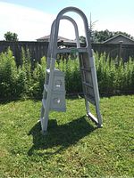 Full view of white plastic above ground pool ladder standing on grass with four steps and curved handle.