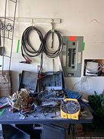 Overview of items on table including electrical cables hung on wall, gloves, clamps, hand tools, extension cord reel, and tool cases
