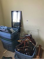 Stack of seven blue and gray Rubbermaid plastic totes with lids on top, leaning full-length mirror, white metal hanging racks in the background, wooden hangers in a blue tote in front.