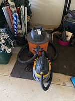 Photo showing both vacuums side by side; large orange and black Rigid vacuum and smaller gray and yellow Stinger vacuum with hoses attached.