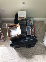 Photo showing Toshiba VHS/DVD player with a cardboard box filled with music CDs, and several stacks of DVDs beside it, all placed on a carpeted floor.