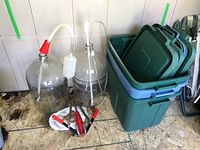 Photo showing two large glass wine carboys, one with a red and white plastic stopper and attached tubing, the other with a white plastic airlock and tubing. In front is a red and silver hand corker and several wine corks on a white piece of cardboard. To the right are several stacked green plastic storage totes with lids.