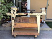 Frontal view of lathe mounted on wooden mobile base with tool storage shelves and swivel casters.