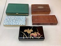 Five vintage jewelry boxes arranged in a group showing a variety of colors and materials including lacquer, leather, wood and fabric.