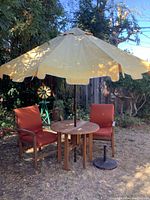 Full view of outdoor patio set including table, umbrella, stand, and two chairs in garden setting with trees and plants in background.